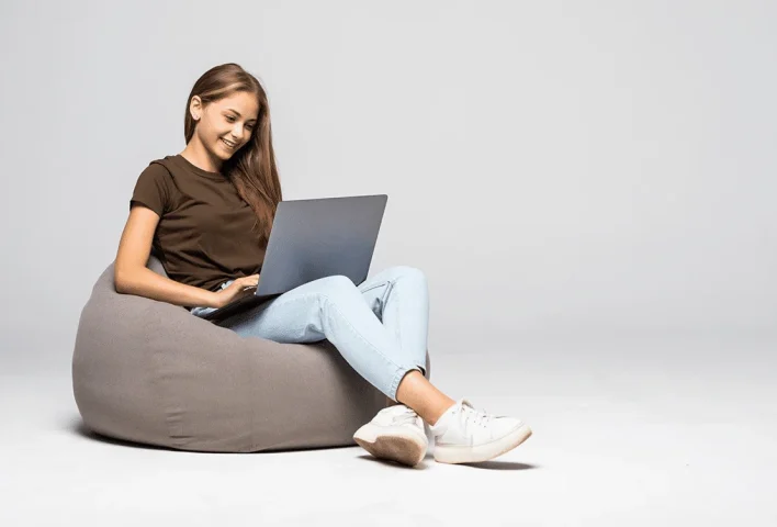 A teenage girl is sitting on a comforter, holding a laptop and wearing white sneakers.